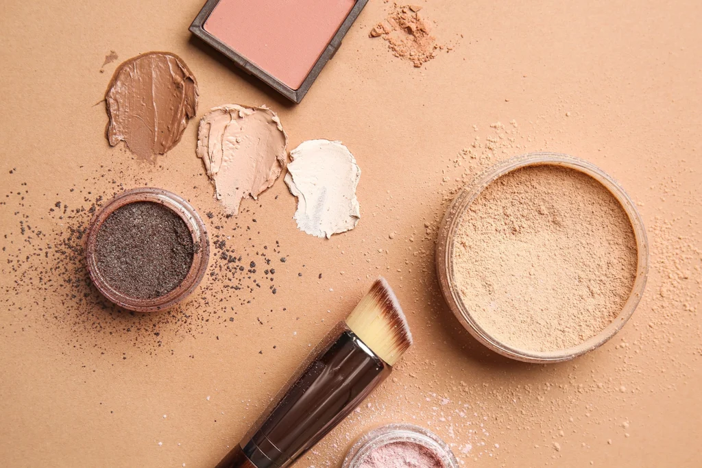 Varie texture di cosmetici in polvere e crema, con un pennello da trucco su sfondo beige, a rappresentare l'importanza di leggere l'INCI dei prodotti cosmetici per scegliere ingredienti sicuri e consapevoli.