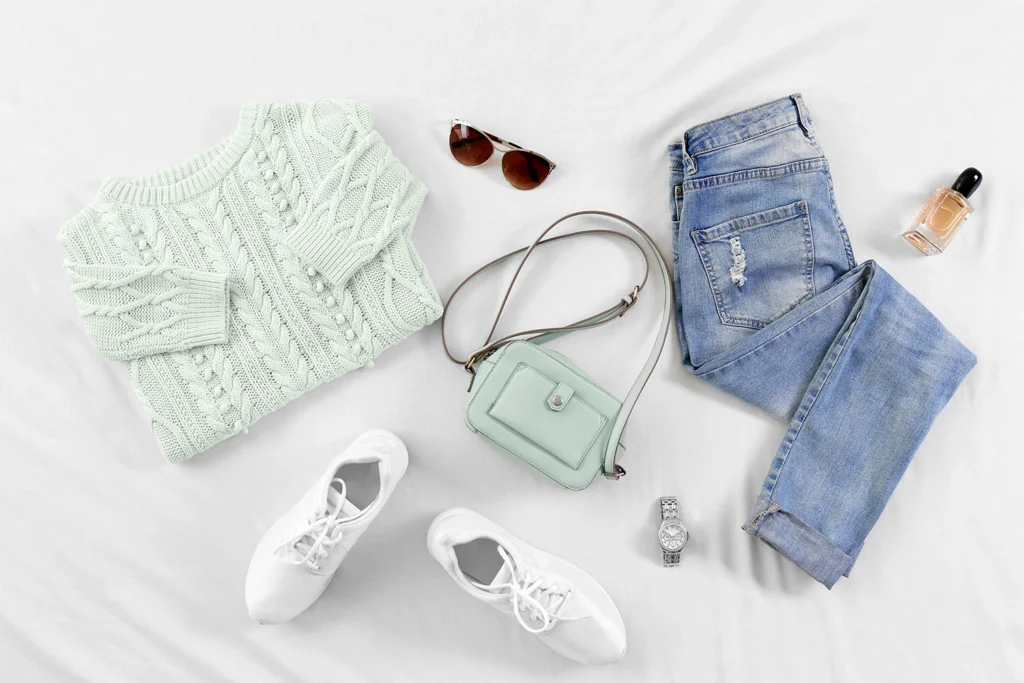 Flat lay di capi essenziali per un armadio capsula: maglione di lana verde chiaro, jeans, sneakers bianche e borsa minimal.