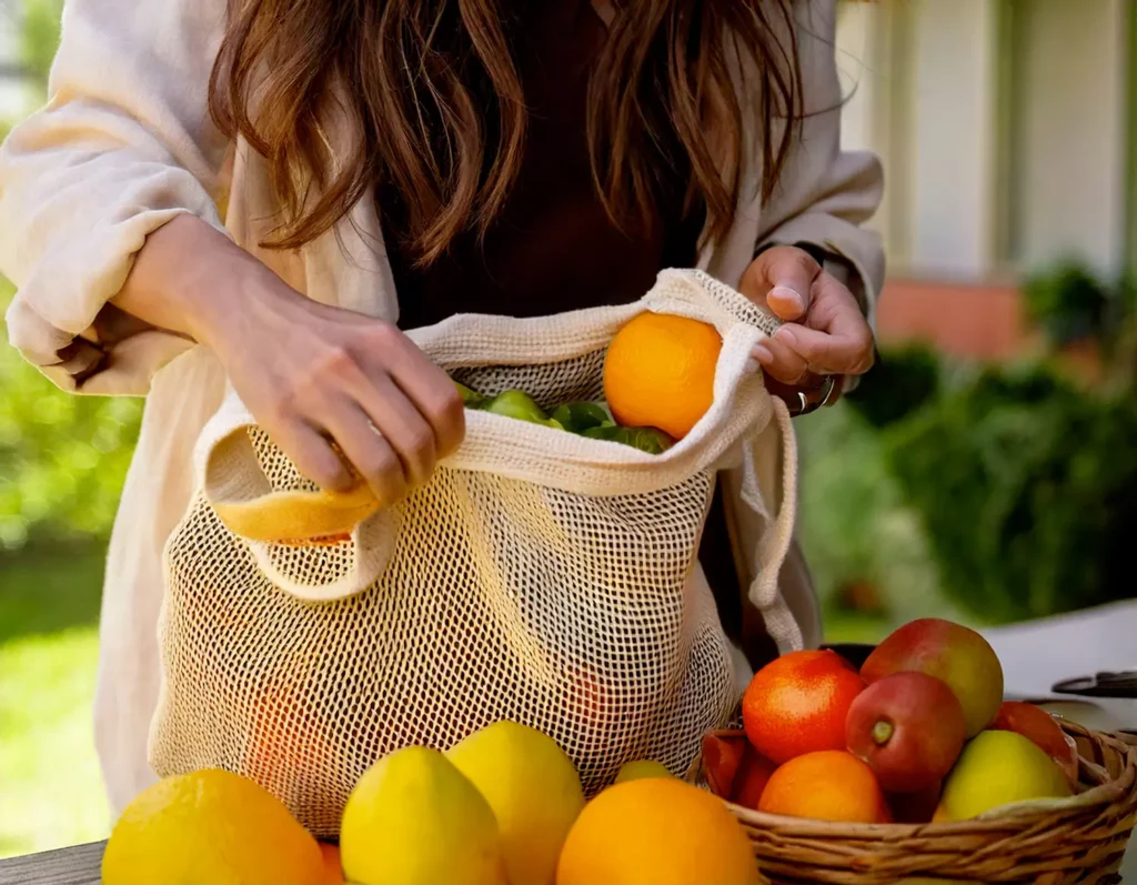 Mani che riempiono un sacchetto in cotone riutilizzabile con frutta e verdura fresca, esempio di materiali sostenibili e alternative eco-friendly per ridurre la plastica nella vita quotidiana.  