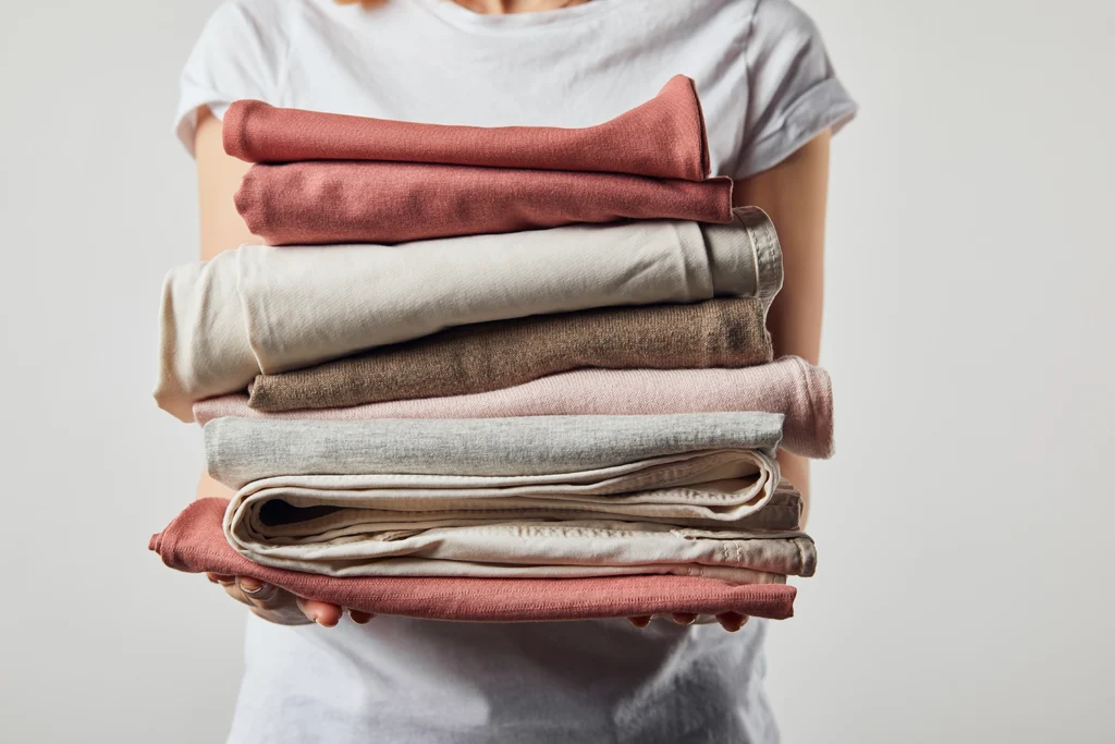 Persona che tiene in mano una pila di capi essenziali in tonalità neutre, esempio pratico di guardaroba minimal e armadio capsula, perfetto per il minimalismo nel guardaroba.