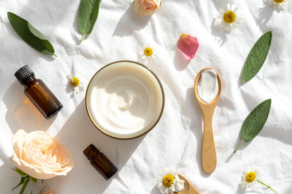 Prodotti skincare vegani per pelle secca con crema, boccette in vetro ambrato, petali di rosa e fiori di camomilla su fondo bianco illuminato dal sole