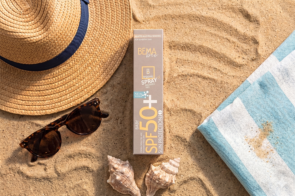 Crema Spray Solare SPF 50+ Baby di Bema Cosmetici nella sua confezione di cartone sulla sabbia con occhiali da sole, un cappello di paglia e due conchiglie