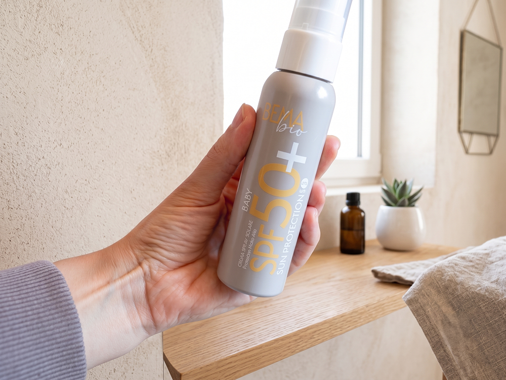 Mano che tiene il flacone di Crema Spray Solare SPF 50+ Baby Bema Cosmetici in un bagno luminoso dal design minimal ed elegante