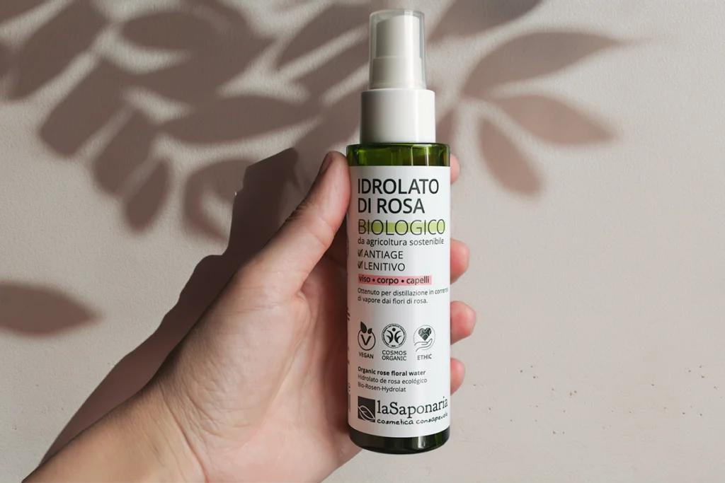 Idrolato di Rosa La Saponaria biologico da agricoltura sostenibile, flacone spray verde tenuto in mano su sfondo neutro con ombre di foglie